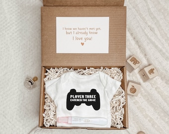 May include: A brown cardboard box with a white onesie inside. The onesie has a black graphic that says "Player Three Entered the Game" with a video game controller icon. The box also contains a pregnancy test, a pacifier, wooden blocks, and a wooden rattle.