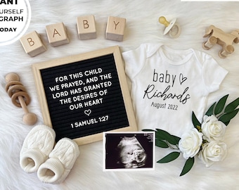 Pode incluir: Um macacão branco com o texto "baby Richards August 2022" nele, um quadro de letras com o texto "For this child we prayed, and the Lord has granted the desires of our heart ♡ 1 Samuel 1:27", blocos de madeira que soletram "BABY", um sapatinho de bebê de tricô branco, um chocalho de madeira, um cavalo de madeira, uma chupeta, uma imagem de ultrassom branca e flores brancas com folhas verdes em uma superfície branca felpuda.