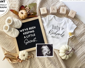 May include: A white onesie with the text "baby Richards March 2022" and a heart symbol. A black letter board with the text "We've been keeping a little secret..." is in the background. There are also baby booties, a pacifier, and a pumpkin.