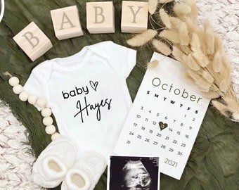 May include: A white onesie with the text "baby Hayes" and a heart symbol.  A calendar with the month of October highlighted with a heart on the 13th.  Wooden blocks spelling out "BABY" and a bouquet of dried pampas grass.