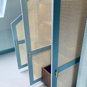 Puede incluir: Tres puertas de armario de color azul claro con paneles de ca&ntilde;a tejida y tiradores de lat&oacute;n. Las puertas est&aacute;n abiertas, revelando un caj&oacute;n de madera en el interior.