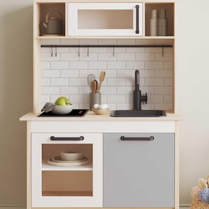 May include: A white and wood children's play kitchen with a black sink, stovetop, and cabinet hardware. The kitchen has a gray cabinet door and a glass-fronted cabinet door with a shelf inside. There are hooks above the sink for hanging utensils.