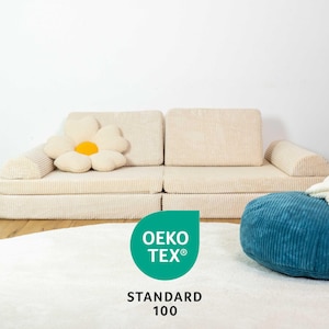 May include: A beige corduroy sofa with a large white flower cushion. The sofa is in front of a white wall. A blue corduroy pouffe sits in front of the sofa. The text "OEKO-TEX&reg; STANDARD 100" is on a green circle in the foreground.