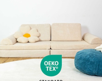 May include: A beige corduroy sofa with a large white flower pillow. The sofa is in front of a white wall. A blue corduroy pouf sits in front of the sofa. The text "OEKO-TEX® STANDARD 100" is on a green circle in the foreground.