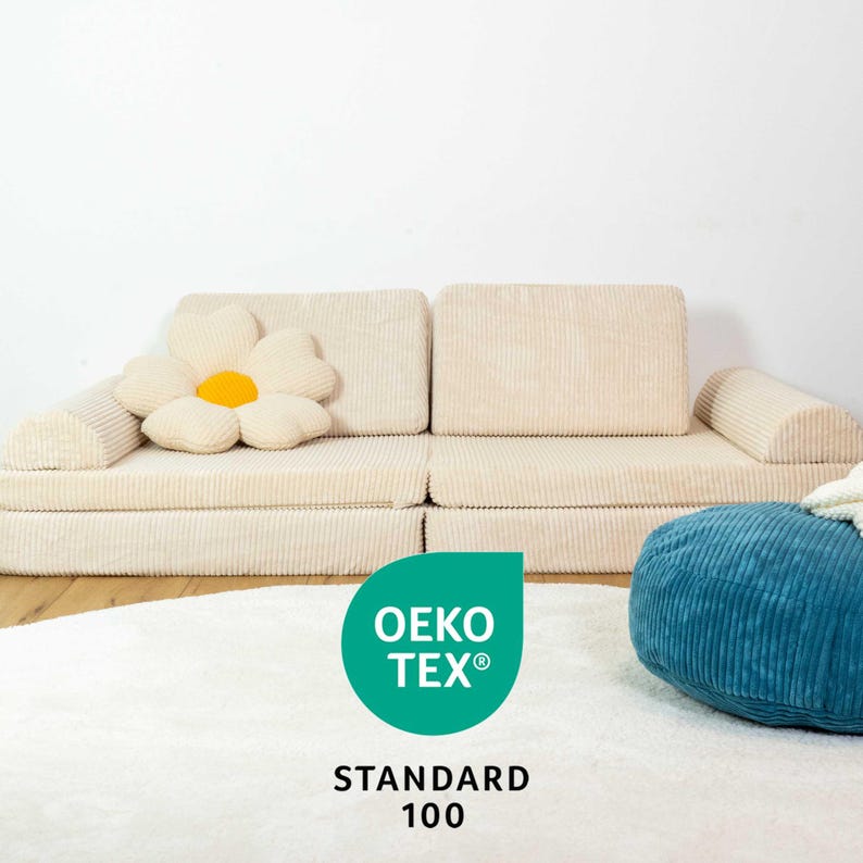 May include: A beige corduroy sofa with a large white flower cushion. The sofa is in front of a white wall. A blue corduroy pouffe sits in front of the sofa. The text "OEKO-TEX&reg; STANDARD 100" is on a green circle in the foreground.