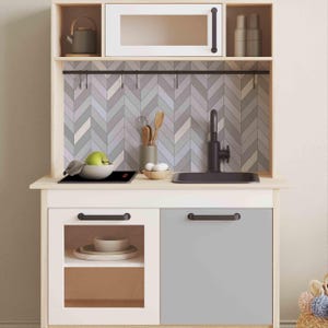 May include: A white and wood play kitchen with a gray sink, black faucet, and a black stovetop. The backsplash is a gray and white chevron pattern. There are hooks above the stovetop for hanging utensils.