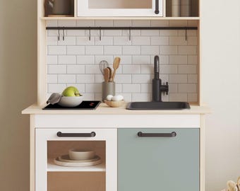 Può includere: Una cucina giocattolo in legno e bianco con un lavello, un piano cottura e maniglie degli armadietti neri. La cucina ha un backsplash in piastrelle bianche e uno scaffale con ganci per appendere gli utensili.