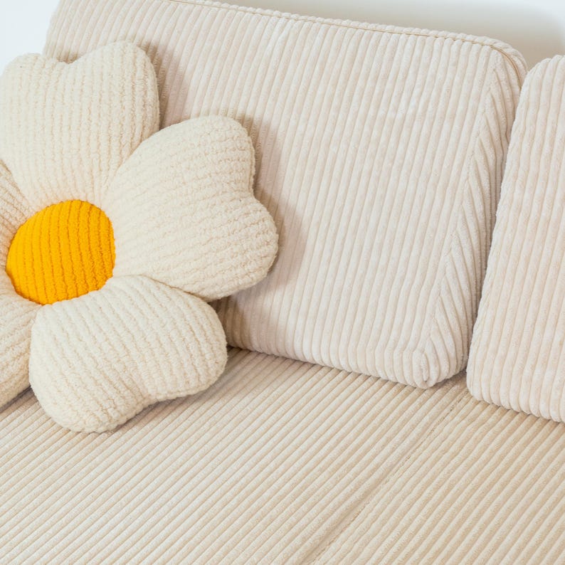 May include: A white corduroy sofa with a large white flower-shaped cushion with a yellow centre. The sofa has a tufted back and a smooth seat.