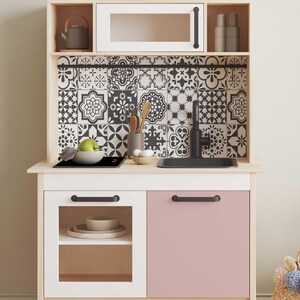 May include: A white and pink play kitchen with a black and white patterned tile backsplash, a black stovetop, a black sink, and a black faucet. The kitchen has two cabinets with black handles and a glass door on the left cabinet.