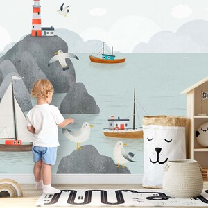 May include: A child stands in front of a wall mural depicting a seascape with a lighthouse, boats, and seagulls. The mural is painted in shades of blue, gray, and white. The child is wearing a white shirt and blue shorts.
