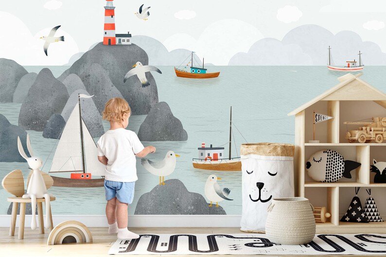 May include: A child stands in front of a wall mural depicting a seascape with a lighthouse, boats, and seagulls. The mural is painted in shades of blue, gray, and white. The child is wearing a white shirt and blue shorts.