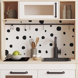 May include: A white play kitchen with black polka dot wallpaper, a black stovetop, a black sink, and a black faucet. The kitchen has a white cabinet with a glass door and a shelf above the sink. There are two black hooks on the cabinet door.