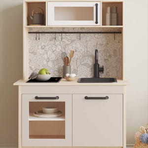 Puede incluir: Una cocina de juguete de madera blanca con un salpicadero de papel tapiz floral negro, un fregadero negro con un grifo, una placa de cocción negra y dos armarios con tiradores negros.