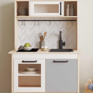 May include: A white and gray play kitchen with a black sink, stovetop, and faucet. The kitchen has a white cabinet with a glass door and a gray cabinet with a solid door. The kitchen has a wooden frame and a white countertop.