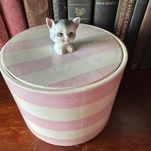May include: A ceramic container with a lid featuring a pink and white striped pattern. A small, detailed figurine of a kitten sits atop the lid. The container is cylindrical and sits on a wooden surface.