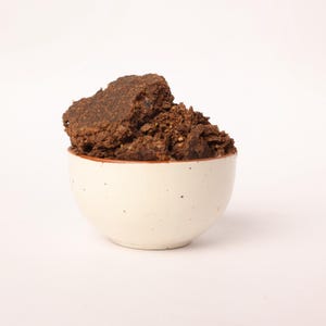 Peut inclure: Un petit bol en céramique blanc cassé, tacheté, rempli d'une grande portion d'aliments brun foncé et friables. Le bol a un fin bord brun rougeâtre. La nourriture semble être une masse solide avec une surface rugueuse et texturée.