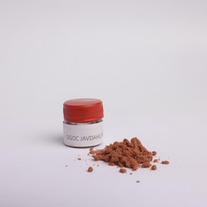May include: A small glass jar with a red lid, labelled "OGOC JAVDAHUS", and a pile of brown powder. The jar and powder are on a white surface. The powder appears to be a fine, granular substance.