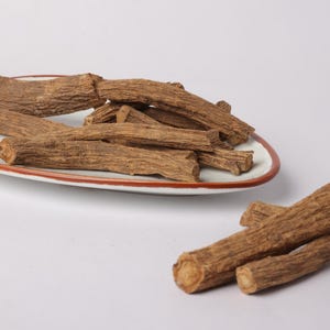May include: A selection of brown, textured, dried roots, some presented on a white plate with an orange rim, others scattered around. The roots have a rough, bark-like texture, suggesting a natural product. The image is set against a plain white background.