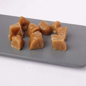 May include: A collection of light brown, irregularly shaped food items, possibly fudge or candy, arranged on a gray rectangular surface. The pieces have a slightly textured appearance and varying sizes, suggesting a homemade or artisanal quality.
