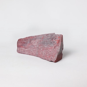 Op de afbeelding: Een ruw bewerkt, rood-paars mineraal specimen met een textuur. De steen heeft een rechthoekige vorm met een gekartelde rand en staat tegen een witte achtergrond. De kleur is een mix van dieprood en lichter paars.