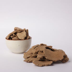 以下が含まれることがあります： 白のセラミックボウルに、形が不規則な薄茶色の破片が詰められ、同様の破片の山が隣にあります。これらのアイテムは、プレーンな白い背景に対して配置されており、すっきりとしたシンプルなプレゼンテーションになっています。