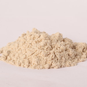 May include: A pile of light beige powder, possibly flour or a similar dry ingredient, sits against a white background. The powder has a fine, granular texture and appears to be freshly milled.