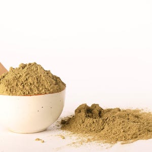 May include: A white ceramic bowl filled with a light brown powder, with a wooden spoon resting inside. A small pile of the same powder sits next to the bowl on a white surface. The powder has a fine texture.