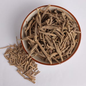 May include: A top-down view of a ceramic bowl filled with dried, beige-colored ashwagandha roots. Some roots are scattered outside the bowl on a white surface. The bowl has a brown rim.