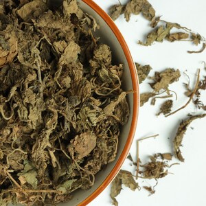 Puede incluir: Primer plano de hojas secas marrones que llenan un cuenco de cerámica con un borde marrón y naranja. Algunas hojas están esparcidas sobre la superficie blanca. Las hojas parecen ser un producto herbal, posiblemente para té o uso medicinal.