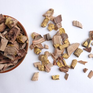 May include: A ceramic bowl containing brown and tan wood chips, with additional chips scattered on a white surface. The wood chips display a variety of textures and colours, including some with a golden tint. The image is well-lit, highlighting the details of the wood.