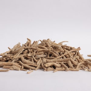 May include: A pile of dried, light brown ashwagandha roots against a white background. The roots are of varying lengths and thicknesses, with a rough, textured appearance. The image is well-lit, highlighting the natural color and form of the herbal product.