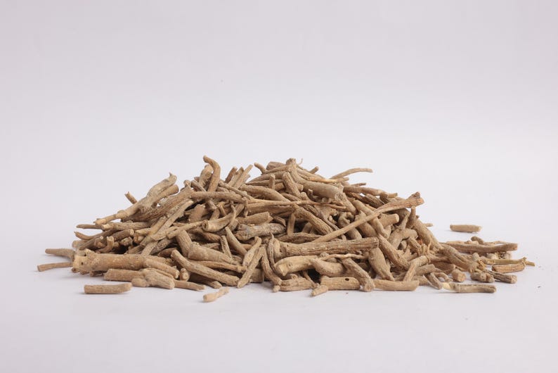 May include: A pile of dried, light brown ashwagandha roots against a white background. The roots are of varying lengths and thicknesses, with a rough, textured appearance. The image is well-lit, highlighting the natural color and form of the herbal product.