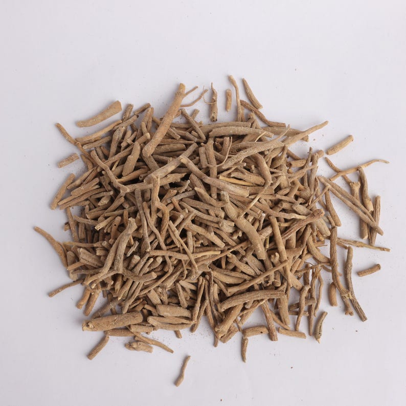 May include: A pile of dried, light brown ashwagandha roots on a white background. The roots are of varying lengths and thicknesses, with a rough, textured appearance. The image is a close-up, showing the details of the herbal product.