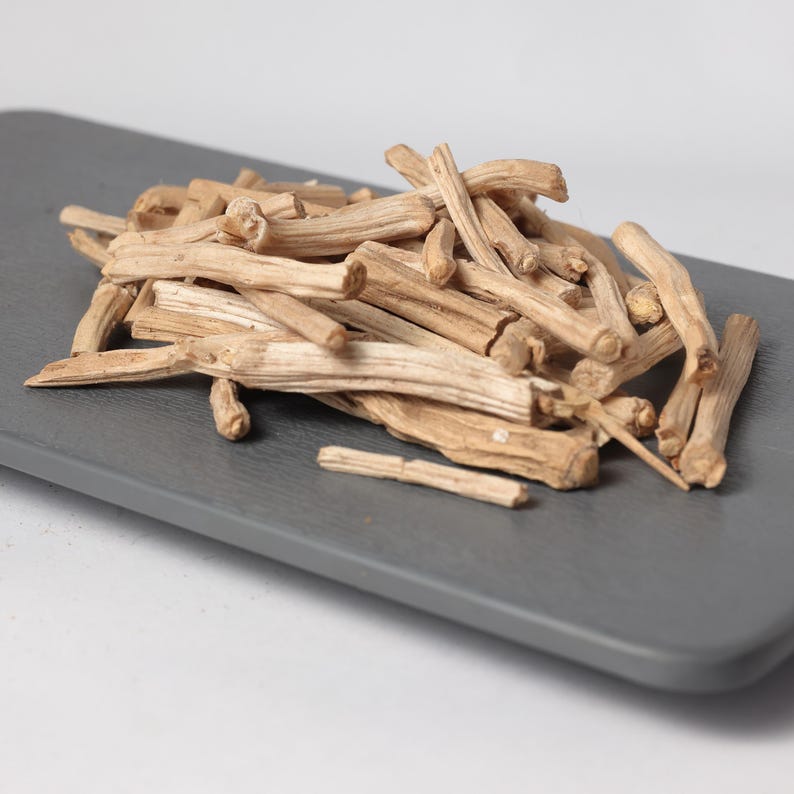 May include: A pile of dried, light brown, woody stems, possibly herbs or spices, arranged on a gray rectangular surface. The stems vary in thickness and length, with a natural, organic appearance. The background is a plain white.