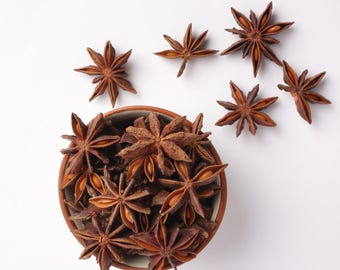 May include: Overhead shot of star anise spices. A small, round, brown bowl is filled with star anise, with several more scattered around it on a white surface. The star anise is a deep brown color and has a star-like shape.