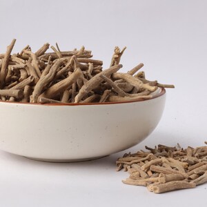 May include: A white ceramic bowl filled with dried, light brown ashwagandha roots. A small pile of the roots sits beside the bowl on a white surface. The bowl has a brown rim.