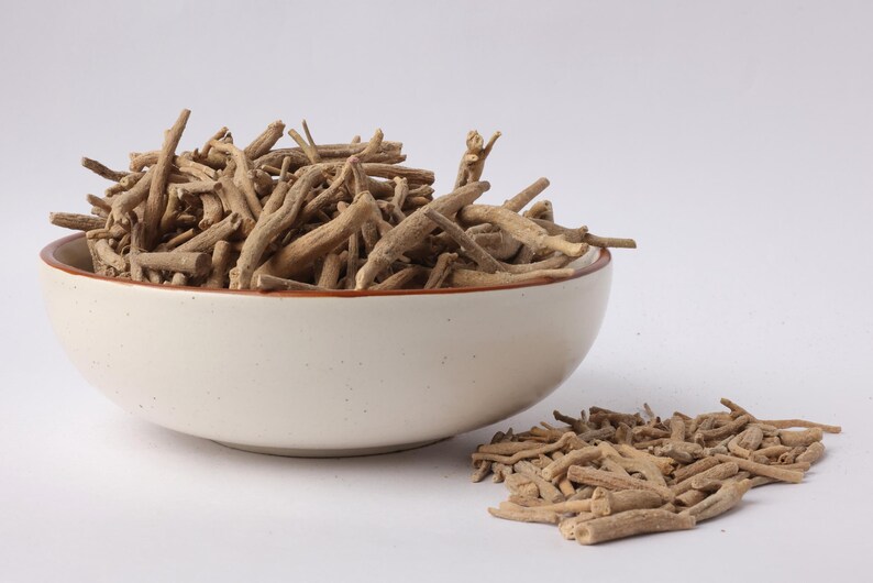 May include: A white ceramic bowl filled with dried, light brown ashwagandha roots. A small pile of the roots sits beside the bowl on a white surface. The bowl has a brown rim.