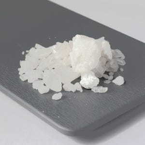 May include: A pile of white, crystalline solids on a gray surface. The crystals vary in size and shape, with some appearing larger and more opaque than others. The background is a solid gray color.