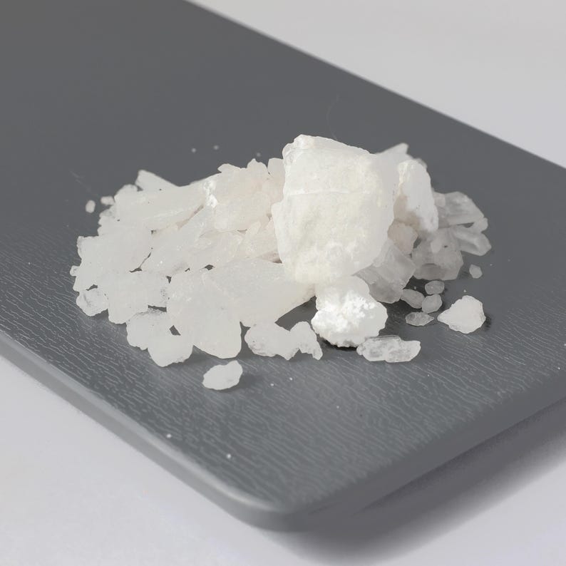 May include: A pile of white, crystalline solids on a gray surface. The crystals vary in size and shape, with some appearing larger and more opaque than others. The background is a solid gray color.
