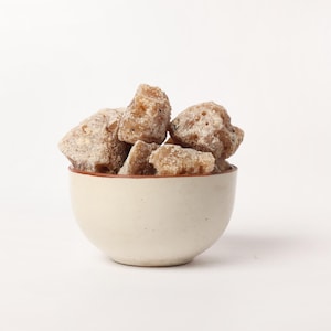 Könnte beinhalten: Eine kleine, cremefarbene Keramikschale gefüllt mit mehreren Stücken braunen, kristallisierten Zuckers. Die Zuckerstücke haben eine raue Textur und sind mit einem feinen, weißen Pulver bedeckt. Die Schale hat ein schlichtes Design mit einem dünnen, braunen Rand.
