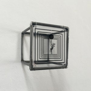 May include: A three-dimensional cube sculpture constructed from dark grey metal framing. A clear acrylic panel is mounted on the front, featuring a black graphic print of concentric squares, a solid black circle, and a hand reaching upward.