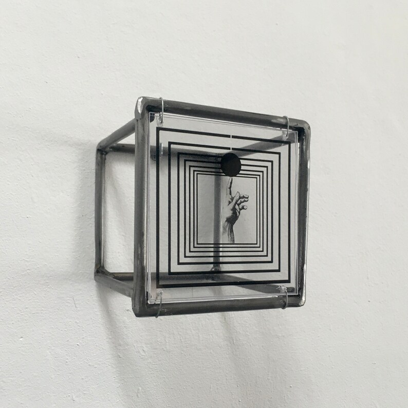 May include: A three-dimensional cube sculpture constructed from dark grey metal framing. A clear acrylic panel is mounted on the front, featuring a black graphic print of concentric squares, a solid black circle, and a hand reaching upward.