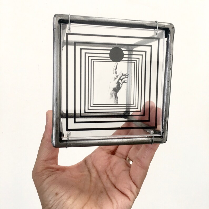 May include: A square, three-dimensional frame constructed from dark, metallic rods. Suspended within the frame is a clear acrylic panel featuring a black graphic print of concentric squares, a solid black circle, and a hand reaching upward.