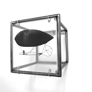 Può includere: Una silhouette in bianco e nero di un dirigibile all'interno di un cubo di vetro con una cornice in metallo.
