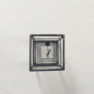 May include: A square, multi-layered glass box frame featuring a black and white graphic print. The print displays a hand with an extended index finger pointing toward a solid black circle, surrounded by a series of concentric black square outlines.