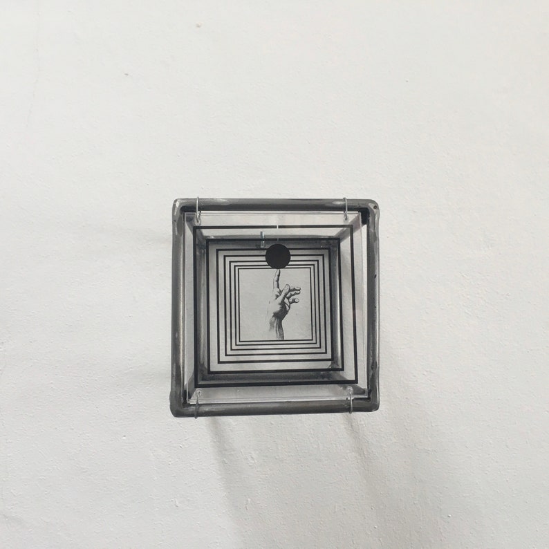 May include: A square, multi-layered glass box frame featuring a black and white graphic print. The print displays a hand with an extended index finger pointing toward a solid black circle, surrounded by a series of concentric black square outlines.