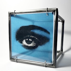 Puede incluir: Una obra de arte enmarcada que presenta un ojo estilizado sobre un fondo azul. El ojo está representado en blanco y negro, con énfasis en la ceja y el párpado superior. El marco es un cubo de metal plateado, con la obra asegurada por pequeños bucles de metal.
