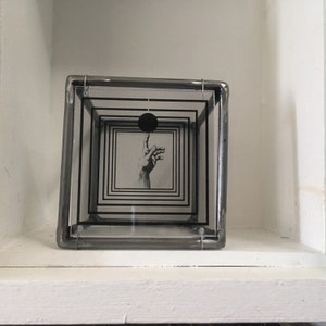 May include: A square glass display box with a dark metal frame containing multiple layered transparent panels. Each panel features a series of concentric black square outlines. A black circle and a detailed illustration of a hand are centered.