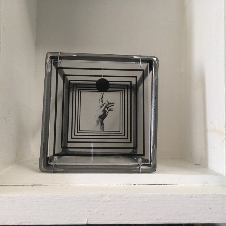 May include: A square glass display box with a dark metal frame containing multiple layered transparent panels. Each panel features a series of concentric black square outlines. A black circle and a detailed illustration of a hand are centered.