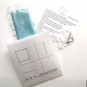 May include: A white envelope with the Art and Shadow logo and geometric line art, a light blue cleaning cloth in a plastic bag, and a small plastic bag containing a metal hook and wall plug, accompanied by an informational instruction sheet.